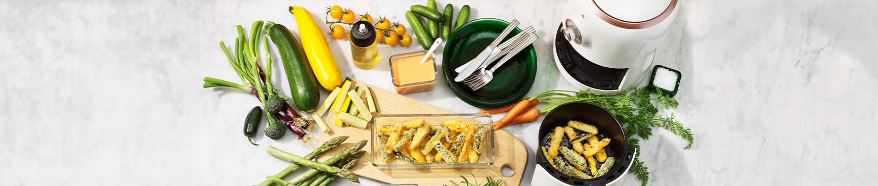 På ett marmorbord står en airfryer och olika sorters grönsaker redo för airfryern, olivolja i en sprayflaska, en skärbräda, tallrikar, bestick och zucchini fries i airfryer.