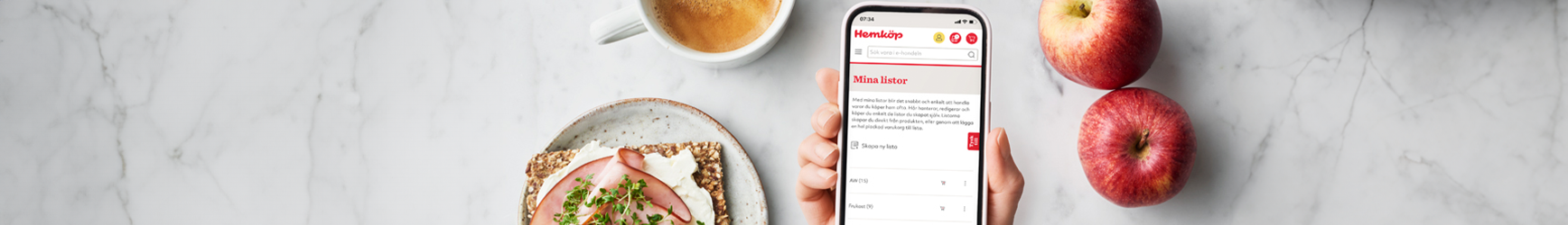 En hand som håller i en mobiltelefon med Hemköp-sidan Mina listor uppe på skärmen. Bakom finns ett marmorbord och en tallrik med en skinksmörgås, två äpplen och en kopp kaffe.