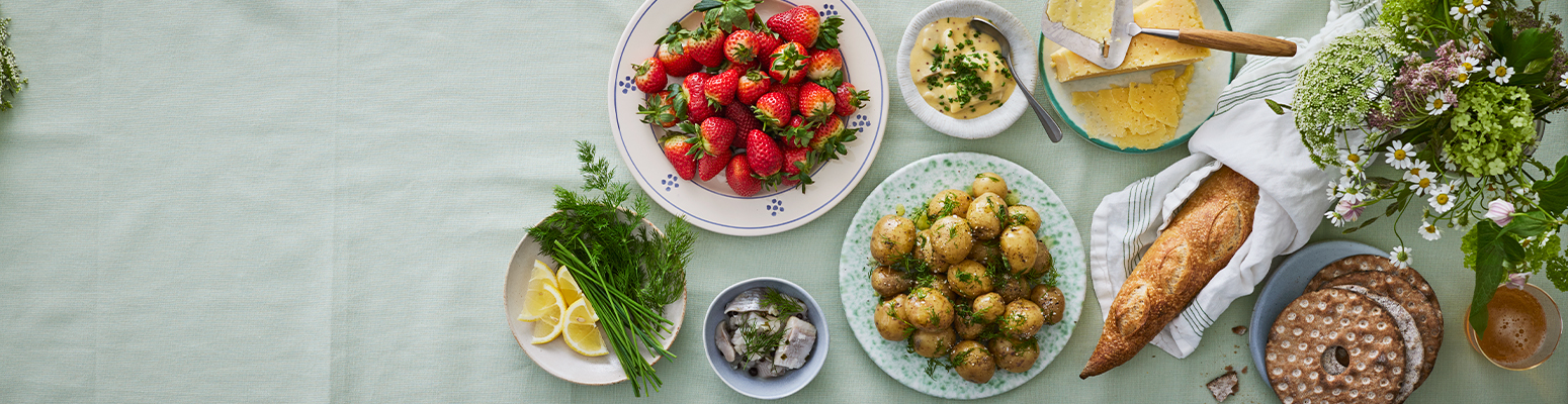 Ett bord med midsommarmat som färskpotatis och dill, jordgubbar, sill, knäckebröd, bröd och ost.