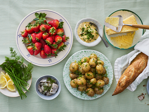 Ett bord med midsommarmat som färskpotatis och dill, jordgubbar, sill, knäckebröd, bröd och ost.