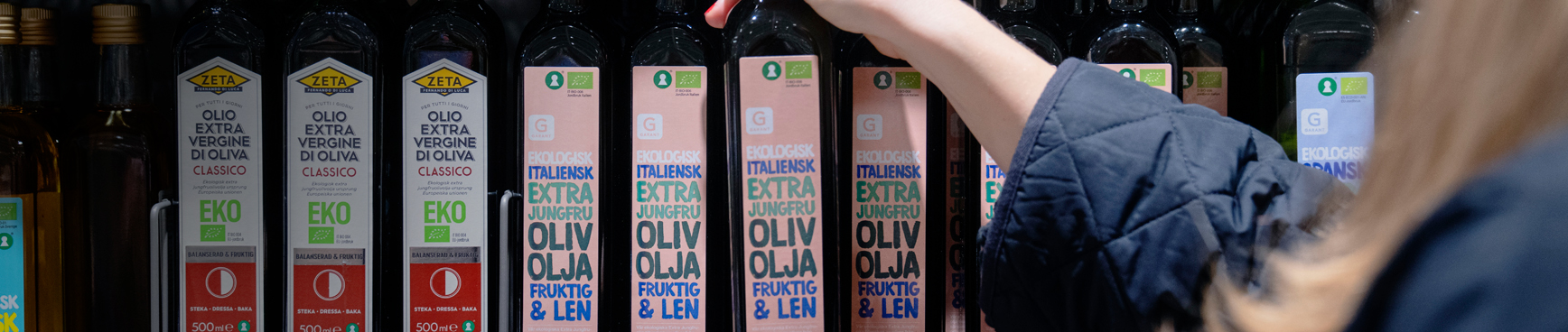 En kvinna tar ner en glasflaska med ekologisk olivolja från en varuhylla i en Hemköpbutik